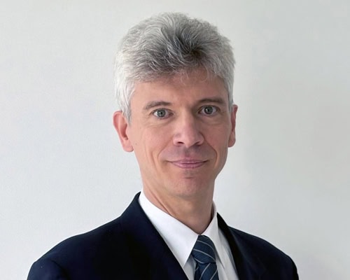 Graue Haare, neutrale Mimik, Anzug mit blauer Krawatte, weißer Hintergrund.