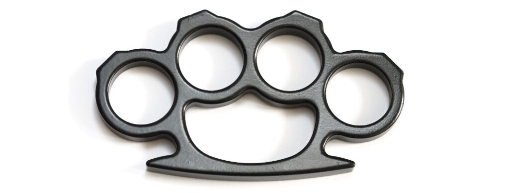 Schwarzer Schlagring aus Metall mit vier Öffnungen auf weißem Hintergrund, ohne erkennbare Beschädigungen.